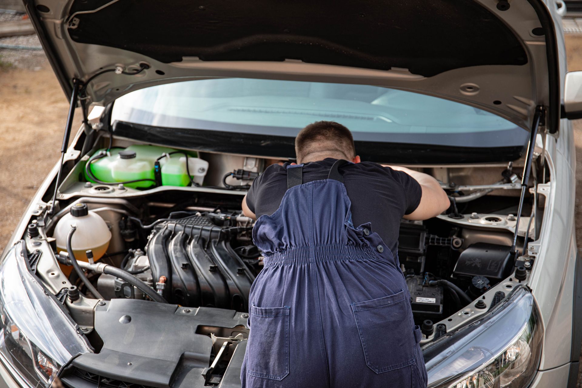 Mechanic in blue overalls working on a car engine outdoors, back view.